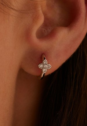 Boucle d’oreille créole en argent avec un motif fleur à quatre pétales ornée de petites pierres précieuses transparentes sur un lobe d’oreille percé.