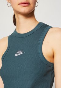 Tyrkysový žebrovaný top bez rukávů s vysokým výstřihem. Na hrudi se nachází malé bílé logo Nike. Materiál má hladkou texturu a přiléhavý střih.