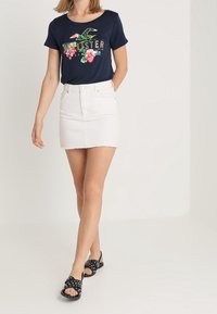 Kvinna som bär en marinblå blommig Hollister-t-shirt, vit denimminikjol och svarta nitar sandaler som går framåt mot en enfärgad bakgrund.