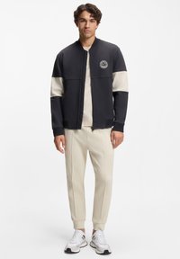 Schwarze und beige Reißverschlussjacke mit rundem Logo, kombiniert mit beigen, zulaufenden Hosen und weißen Sneakers. Verfügt über einen hohen Kragen und gerippte Bündchen.