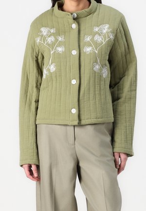 Veste matelassée verte avec broderie florale blanche, dotée d'un col mandarin et de cinq boutons. Associée à un large pantalon beige.