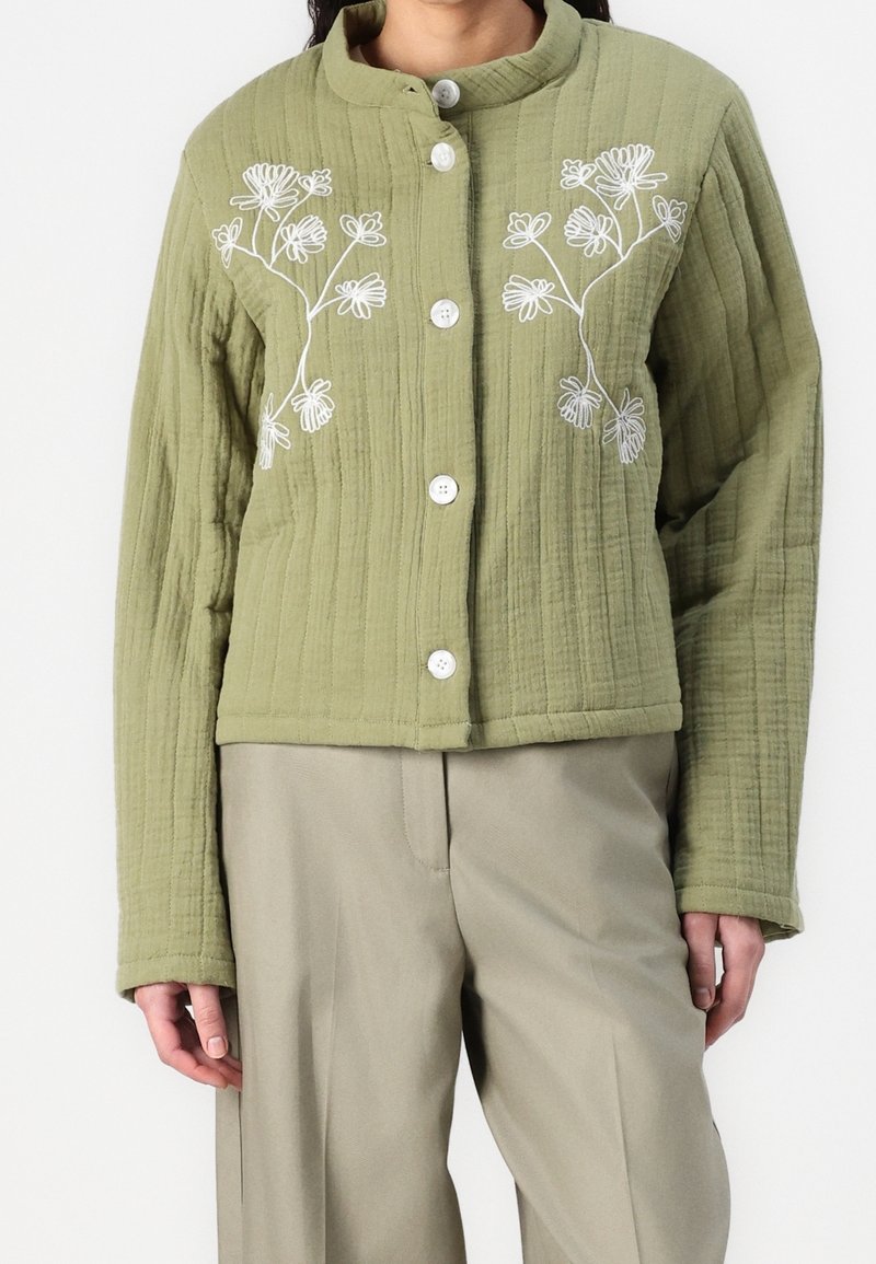 Veste matelassée verte avec broderie florale blanche, dotée d'un col mandarin et de cinq boutons. Associée à un large pantalon beige.
