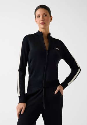 Frau trägt eine schwarze Jacke mit Reißverschluss und weißen Streifen an den Ärmeln sowie passende schwarze Hosen und steht mit einer Hand in der Tasche.