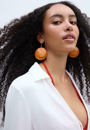 Femme aux cheveux bouclés portant de grandes boucles d'oreilles rondes orange, une chemise blanche et un haut rouge en dessous, regardant avec confiance vers le haut.