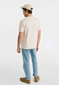 Jasno beżowa koszulka z krótkim rękawem, podwinięte jasnoniebieskie dżinsy i tanowe trampki. Widok z tyłu pokazuje prosty design i luźny krój.