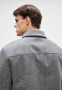 Grau melierte Wollblendenjacke mit klassischem Kragen, sichtbaren Nähten und einem kleinen schwarzen Calvin Klein Logoetikett am hinteren Halsausschnitt.