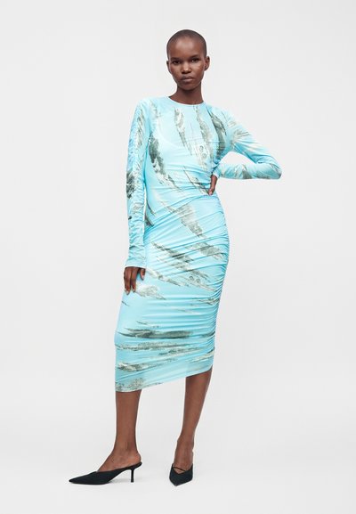 Femme portant une robe midi bleu clair à manches longues avec des motifs abstraits gris, des talons mule noirs pointus, debout avec une main sur la hanche.