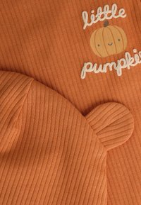 Haut en tissu côtelé orange avec le texte "petit citrouille" et un petit graphique de citrouille ; le chapeau assorti a des oreilles arrondies et une texture côtelée.