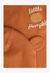 Top de tela acanalada naranja con el texto "pequeña calabaza" y un gráfico de calabaza pequeña; el gorro a juego tiene orejas redondeadas y textura acanalada.
