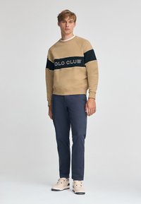 Felpa beige con una fascia nera recante la scritta "POLO CLUB" in lettering bianco, abbinata a pantaloni blu e sneakers chiare.