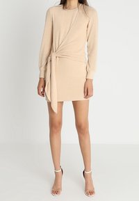 Robe en tricot beige avec des manches longues, design enveloppant et nœud sur le côté. Texture lisse, longueur mi-cuisse, accompagnée de sandales à talons hauts blanches.