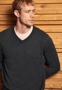 Grau gestrickter V-Ausschnitt-Pullover aus einem strukturierten Material, über ein helles T-Shirt geschichtet, mit einem dezenten Logo auf der linken Brust.