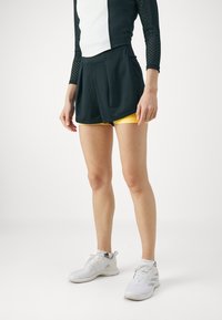 Svart sporttopp med långa, perforerade ärmar, i kombination med svarta shorts och gula underställ. Vita sportskor med svarta detaljer.