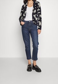 Kvinna som bär avklippta blå jeans, svarta kraftiga loafers, vit linne och en svart-grå geometriskt mönstrad cardigan, står med händerna i fickorna.