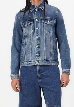 Personne portant une veste en jean bleu à lavage moyen avec fermeture à boutons et deux poches poitrine, associée à un short en jean bleu assorti.