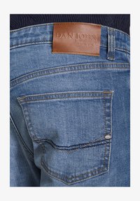 Jeans de mezclilla azul con un parche rectangular de cuero marrón etiquetado "DAN JOHN" en la parte trasera de la cintura y detalles de costura amarilla en los bolsillos.