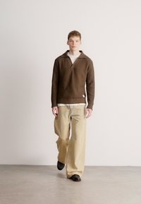 Barna bordázott pulóver, bevágott nyakkal, fehér ingre rétegezve, bézs színű bőszárú nadrággal és fekete cipővel párosítva. Minimalista háttér.