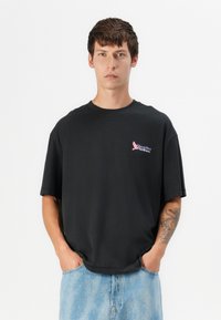 Zwarte oversized t-shirt van katoen, met een klein roze grafisch logo op de linkerkant van de borst. Gepaard met lichtblauwe denim shorts.