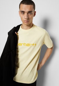 T-shirt de algodão amarelo com decote redondo e mangas curtas, apresentando um logótipo laranja em destaque. O modelo está a usar um casaco preto por um ombro.