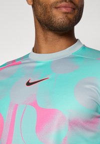 Kortärmad träningsskjorta i ljusblått med rosa och gr å abstrakta mönster. Har en svart Nike-logga på bröstet. Slät yta.