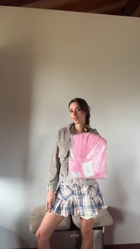Gray corduroy jacket over a white top, paired with a plaid skirt. Holding a pink packaging with a patterned design. Simple background.