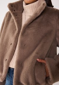 Veste en fausse fourrure de couleur brun clair, avec une texture douce, un col, une fermeture à boutons-pression et des poches latérales. Portée par-dessus un col roulé crème.