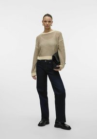 Beige, gestreepte gebreide trui met een losse pasvorm, gecombineerd met donkerblauwe rechte jeans. Model houdt een zwarte clutch vast. Zwarte schoenen maken de look af.