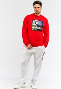 Rød sweatshirt med et grafisk tryk, der indeholder blandet tekst i blå, grøn og sort, parret med hvide bukser og hvide sneakers.