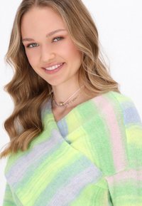 Giovane donna sorridente con capelli mossi indossa un maglione colorato a righe. Collane delicate adornano il suo collo, su uno sfondo bianco semplice.