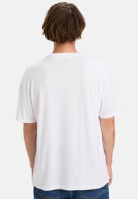 Persoon met kort bruin haar, gekleed in een eenvoudig wit T-shirt met korte mouwen en blauwe jeans, gezien van achteren tegen een witte achtergrond.