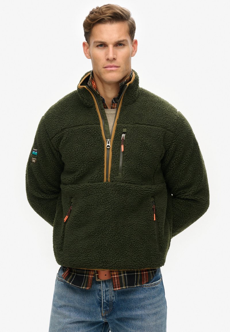 Grüner Fleece-Pullover mit hohem Kragen, Viertelreißverschluss und strukturiertem Oberflächenmaterial. Verfügt über zwei Reißverschlusstaschen und kontrastierenden beige Farbverlauf.