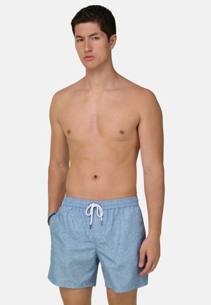 Hombre sin camiseta con pantalones de baño azul claro con estampado y cordón blanco, de pie con una mano en el bolsillo contra un fondo liso.