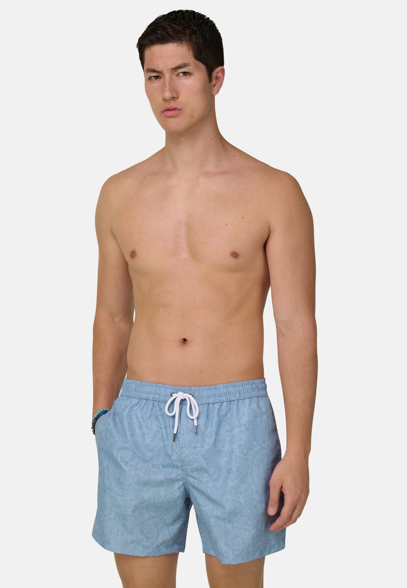 Homme torse nu portant un short de bain bleu clair à motifs avec un cordon blanc, debout avec une main dans la poche sur un fond uni.