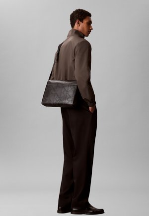 Jonge man draagt een donkerbruine jas en broek, draagt een zwarte geperste leren crossbodytas met een patroon.