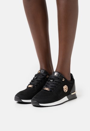 Nohy v černých pletených teniskách s černými tkaničkami, růžově zlatými logy a bílými podrážkami na jednoduchém světlém pozadí.