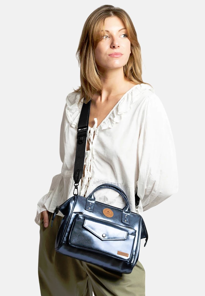 Jeune femme portant un chemisier blanc et un pantalon vert, portant un sac à main bleu foncé brillant avec une bandoulière noire sur l'épaule.