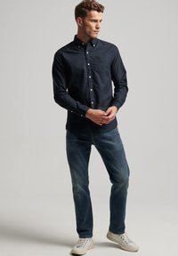 Navy blue button-up shirt with long sleeves, pocket on the chest, paired with light blue jeans and white sneakers.