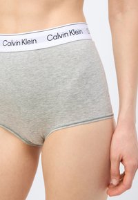 Γυναίκα που φοράει γκρι βαμβακερά εσώρουχα με ψηλή μέση και λευκή ελαστική μέση με τη μάρκα "Calvin Klein" σε απλό φόντο.