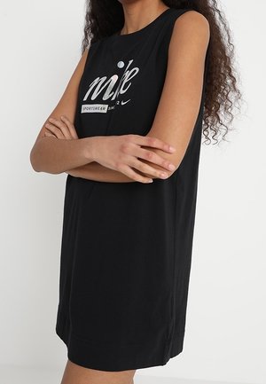 Kobieta z założonymi skrzyżowanymi ramionami, ubrana w bezrękawnikową czarną sukienkę sportową Nike z logo na piersi, stojąca na jednolitym tle.