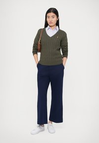 Olivově zelený svetr na kabel s výstřihem do V, v kombinaci s tmavě modrými širokými kalhotami a bílými teniskami. Modelka drží malou hnědou kabelku přes rameno.