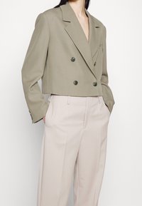 Zkrácená dvouřadá sako v světlém béžovém materiálu s hladkou texturou, vybavené dvěma velkými knoflíky, klopami a tailoring střihem.