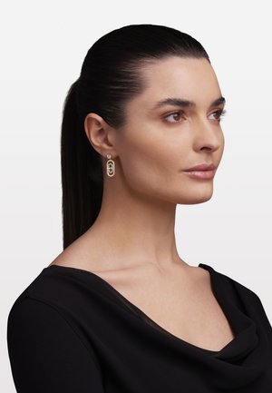 Tumejuustega naine, kes kannab musta tippu ja kuldseid ristkülikukujulisi ripptõkkeid väikese vääriskiviga detailiga.