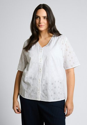 Kobieta z długimi ciemnymi włosami, ubrana w białą bluzkę z oczkami i guzikiem oraz ciemne spodnie w cienkie prążki, stojąca na jednolitym szarym tle.