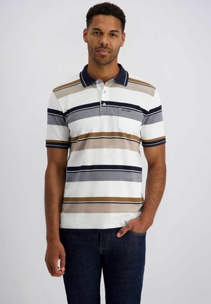 Man die een polo met witte, beige, marineblauwe en grijze strepen draagt, met een donkere spijkerbroek, staand met één hand in de zak tegen een effen achtergrond.