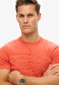 Short-sleeve, round-neck, coral t-shirt with embossed brand text. Features a fitted design. Golden watch with a blue dial on wrist.