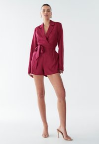 Burgundský volný overal z hladkej tkaniny s dlhými rukávmi, opaskom na viazanie a bočnými vreckami. Má klopový golier a prispôsobený strih.