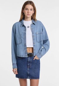 Giacca di jeans blu chiaro con due tasche frontali, chiusura con bottoni e vestibilità morbida, indossata sopra una t-shirt bianca corta e una gonna di jeans.