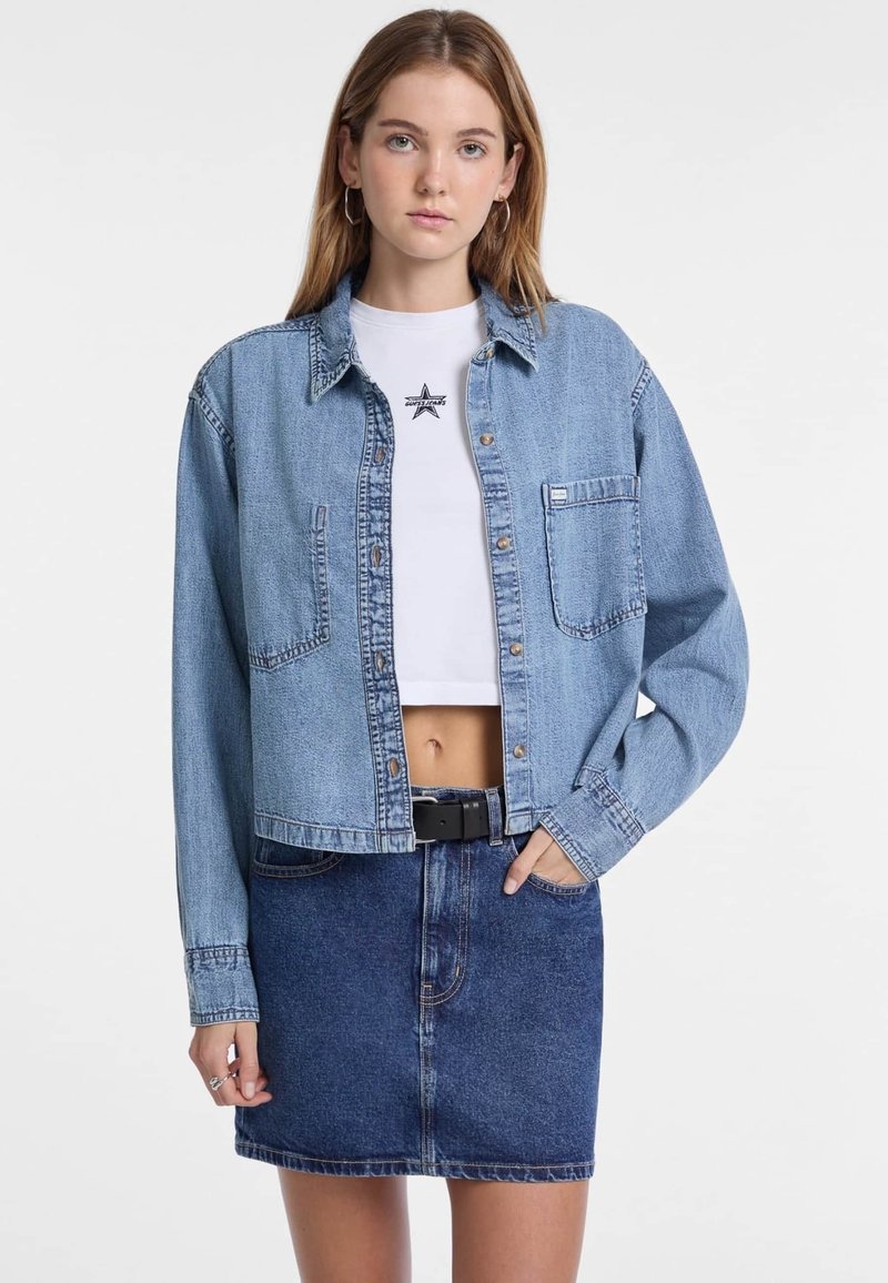 Giacca di jeans blu chiaro con due tasche frontali, chiusura con bottoni e vestibilità morbida, indossata sopra una t-shirt bianca corta e una gonna di jeans.