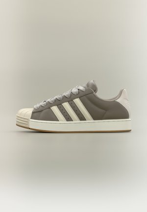 Zapatilla deportiva de cuero gris con acentos blancos, puntera de goma, tres rayas blancas en el lateral y cordones grises. Suela plana con suela exterior de color marrón claro.