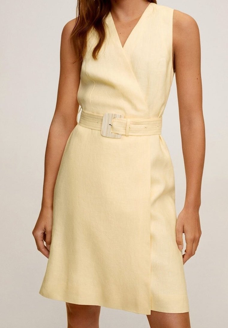 Robe portefeuille sans manches jaune pâle avec une large ceinture assortie et une boucle rectangulaire, portée par une personne debout.